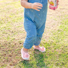 A baby in a denim jumpsuit and pink flower shoes stands on grass, holding a juice box.