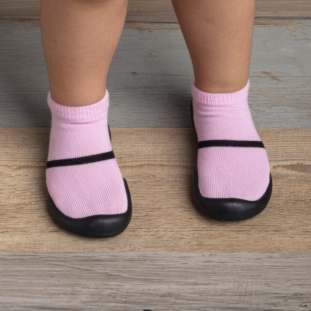 A toddler wearing pink socks with black straps and black rubber soles standing on a wood-grain floor.