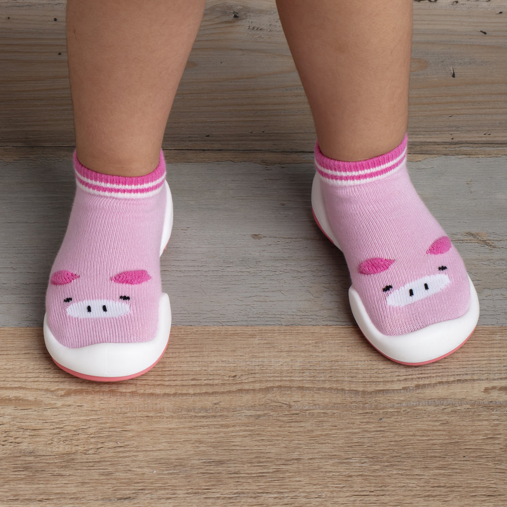 Two feet wearing pink sock-shoes with pig faces on a wooden floor.