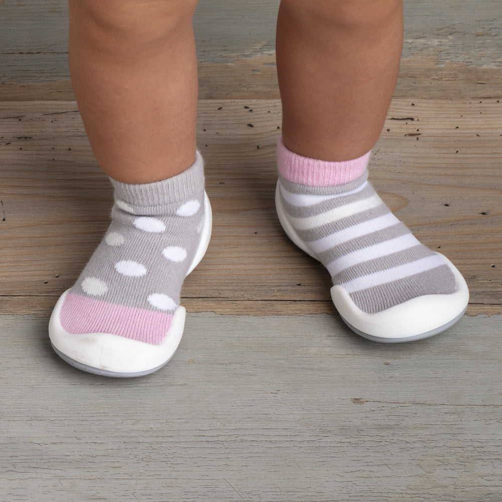 Baby feet wearing gray and white polka dot and striped sock shoes on a wood floor.