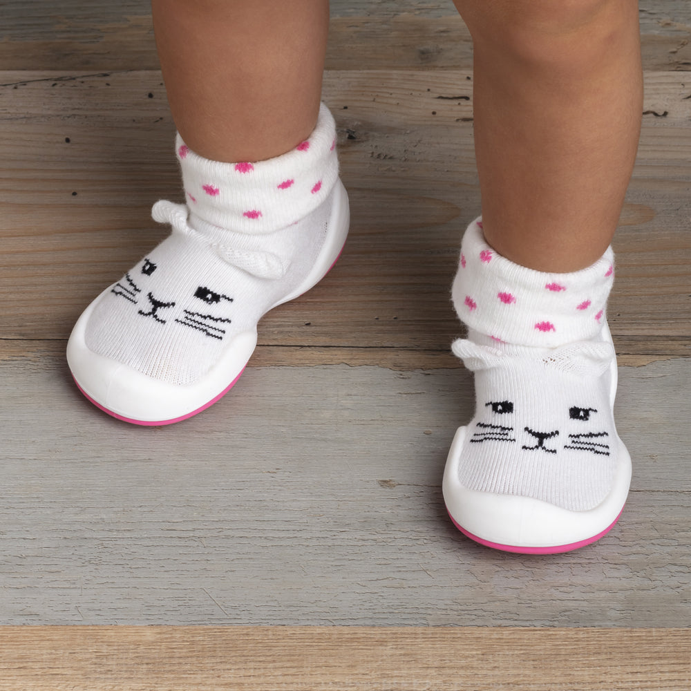 Child's legs and feet wearing white cat-themed shoes with pink polka-dot cuffs on a wooden floor.