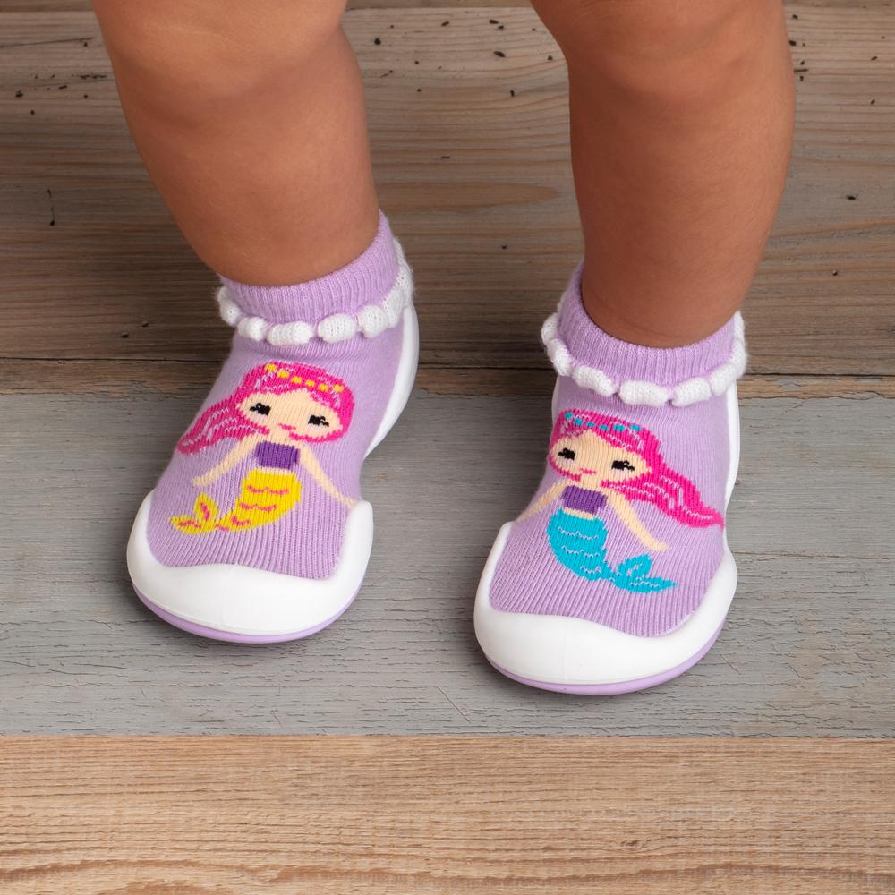 Baby feet wearing purple mermaid-themed shoe socks on a gray wood floor.