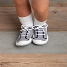 Baby wearing gray knit shoes with white laces and white socks, standing on a wooden floor.