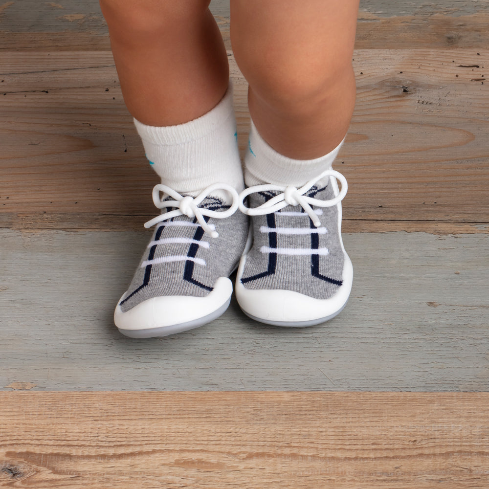 Baby's legs and feet wearing white socks and gray and white sneaker-style slippers on a wooden floor.