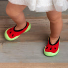A baby wearing watermelon-themed socks and a white and gold dress stands on a wooden floor.