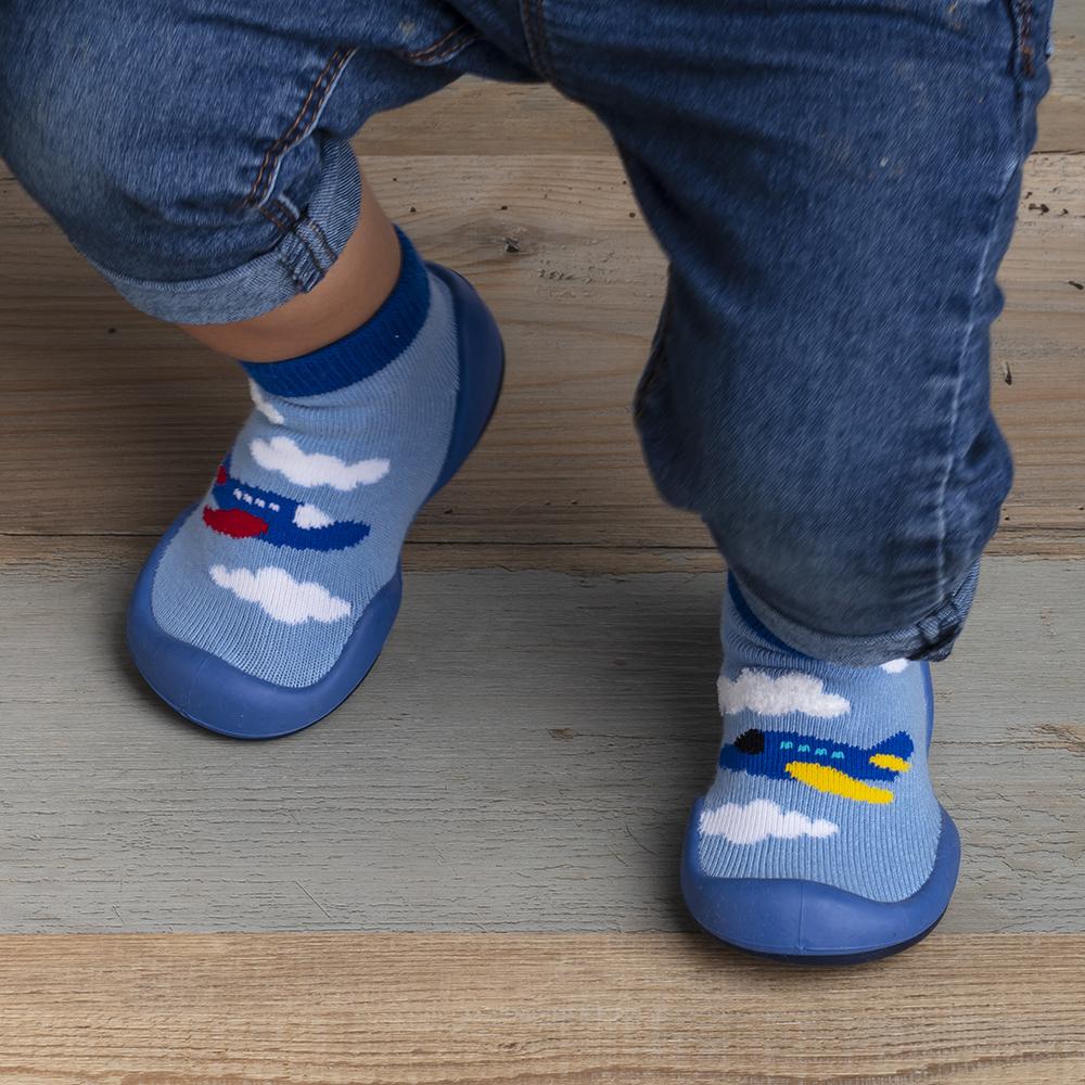 Toddler in jeans wearing blue airplane-themed slipper socks on a wood floor.