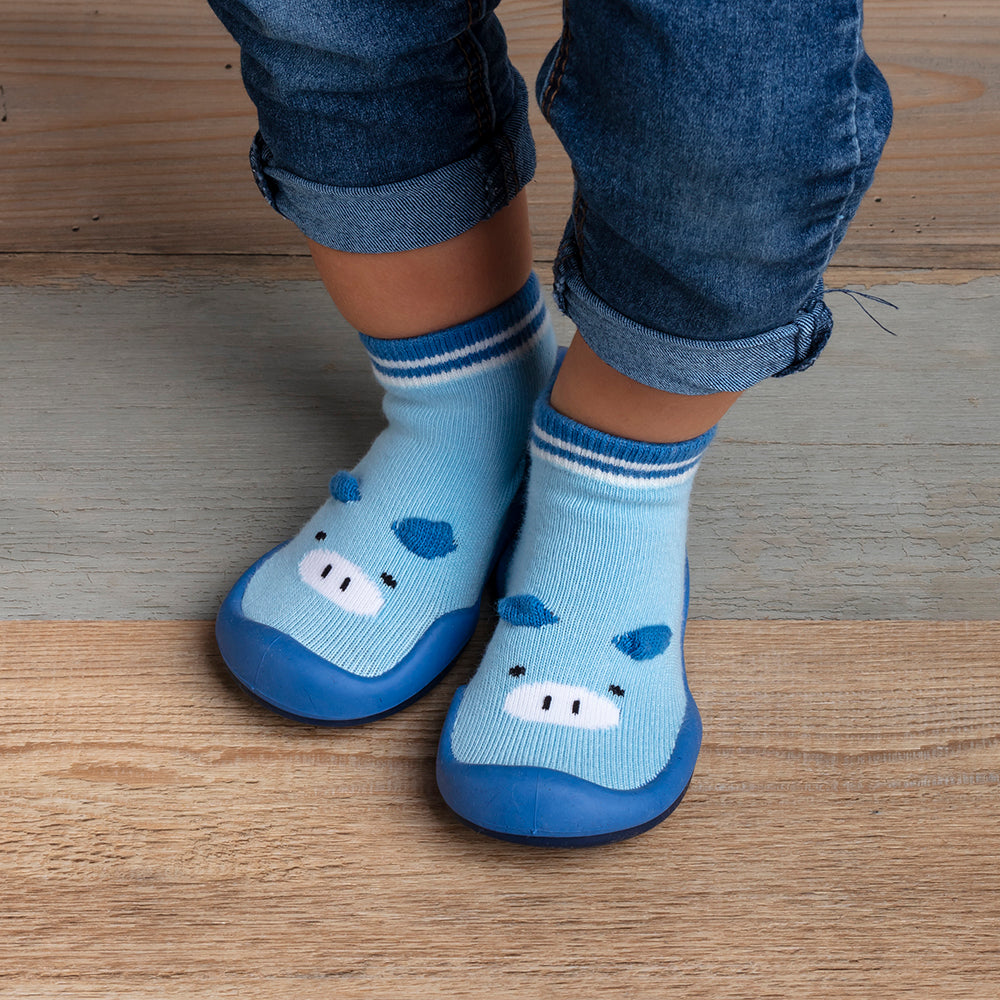 Child wearing blue pig-themed sock shoes with blue jeans on a wood floor.
