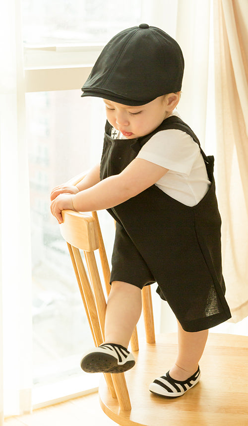 Infant wearing a black cap and overalls with a white T-shirt, standing on a wooden chair in black and white striped shoes.