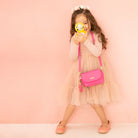 Girl in a pink dress and matching shoes holds a yellow toy with a pink purse against a pink wall.