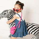 A young girl in heart-shaped sunglasses and a denim dress with a pink purse hugs a zebra toy.
