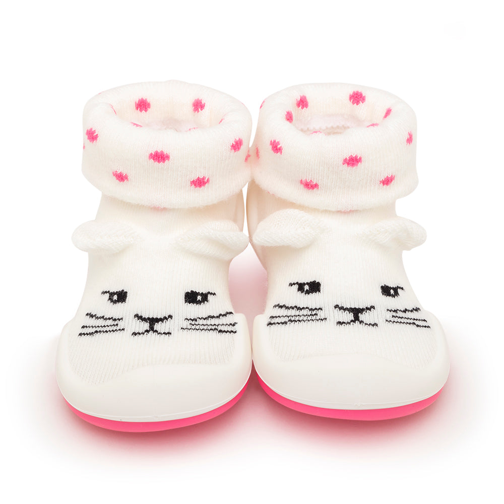 A pair of white baby booties with pink polka dots and a cartoon cat face design, sitting on a white background.