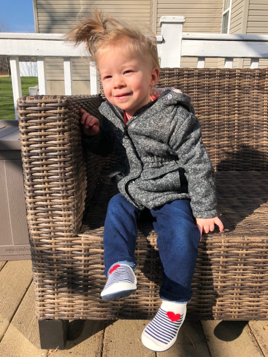 A child in a gray jacket and blue jeans sits on a wicker chair on a sunny day.