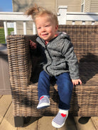 A child in a gray jacket and blue jeans sits on a wicker chair on a sunny day.