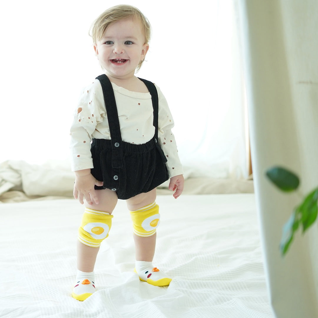 A baby in a white shirt, black overalls, yellow knee pads, and chick-shaped shoes stands on a white bed.