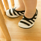 Infant feet in black and white striped shoes stand on wooden floor near chair leg.