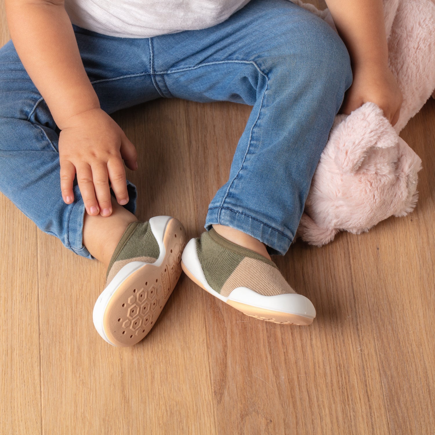 Toddler in jeans and shoes holding a pink stuffed animal, sitting on a wooden floor.