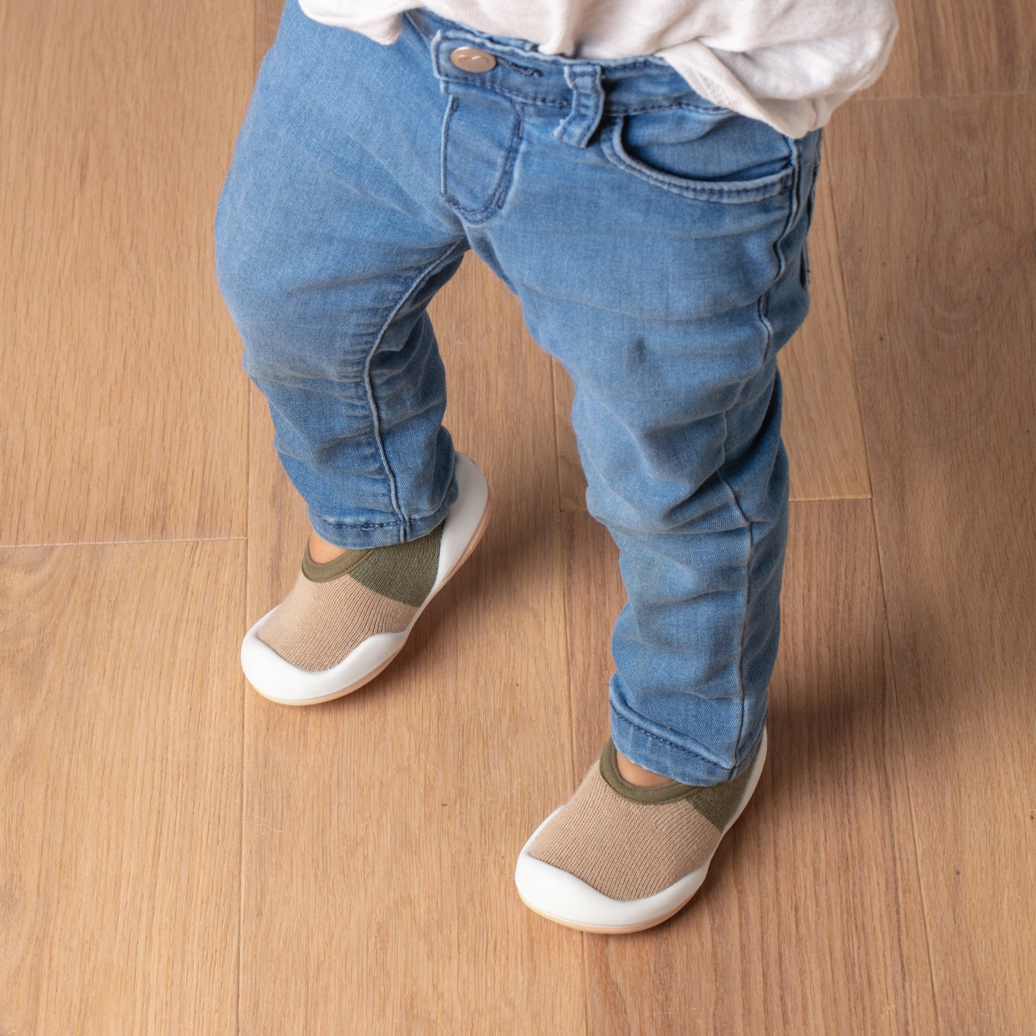 Child's legs in blue jeans and khaki and white shoes, standing on a wood floor.