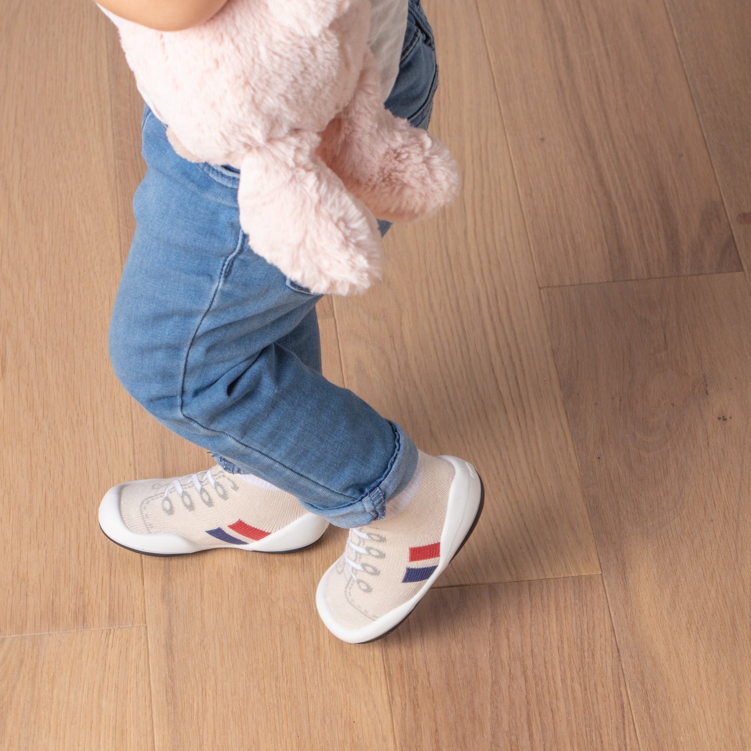 A child wearing jeans and sneaker-socks holds a pink stuffed animal.