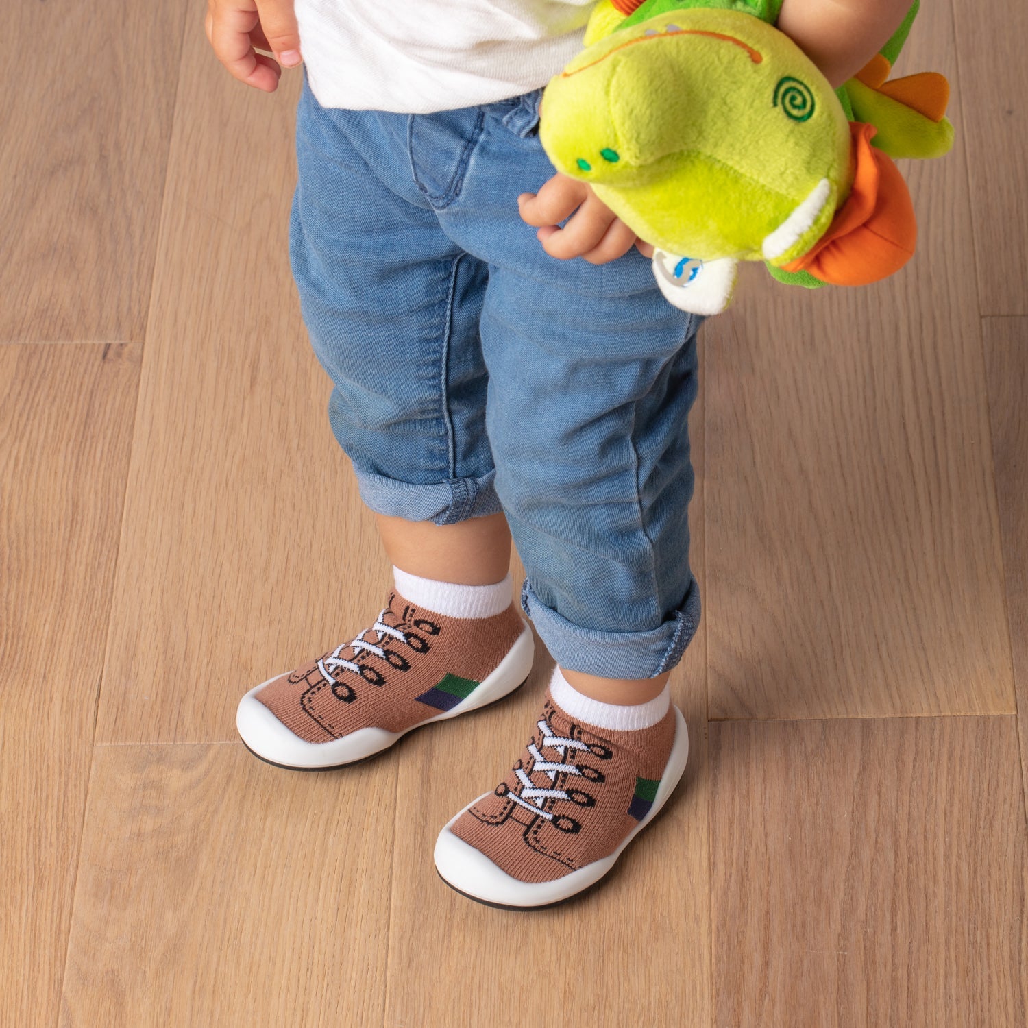 A child with blue jeans and sock shoes, holding a green plush toy.