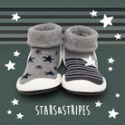 Two baby slipper-socks with white soles, gray fold-down cuffs, and star and stripe designs against a striped green backdrop with white stars and white script.