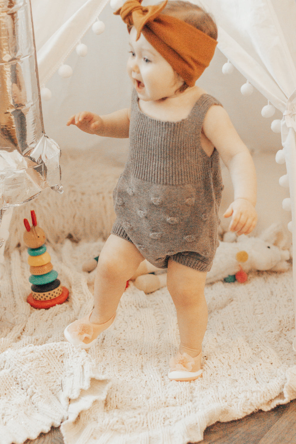 Baby in gray romper with brown bow headband and peach shoes stepping forward on a cream blanket with a wooden stack toy and white stuffed animal nearby.