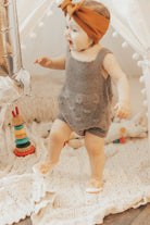 Baby in gray romper with brown bow headband and peach shoes stepping forward on a cream blanket with a wooden stack toy and white stuffed animal nearby.