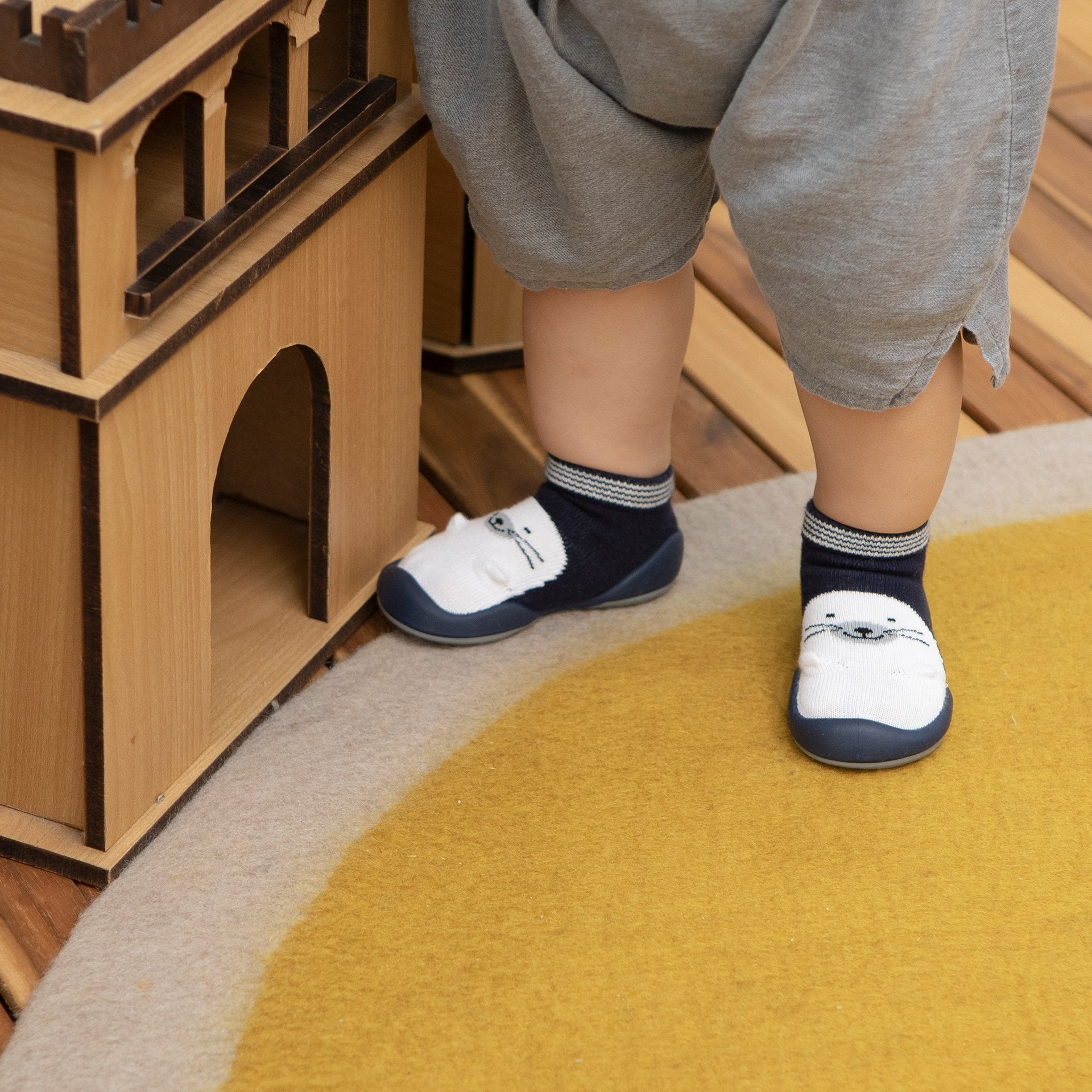 Baby's legs and feet in socks with seal design, gray shorts, wooden castle toy, yellow and beige rug.