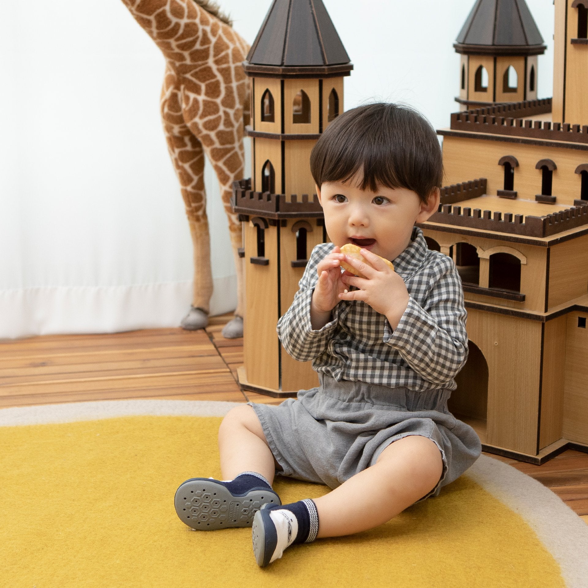 Baby eating on yellow rug next to toy castle and giraffe.