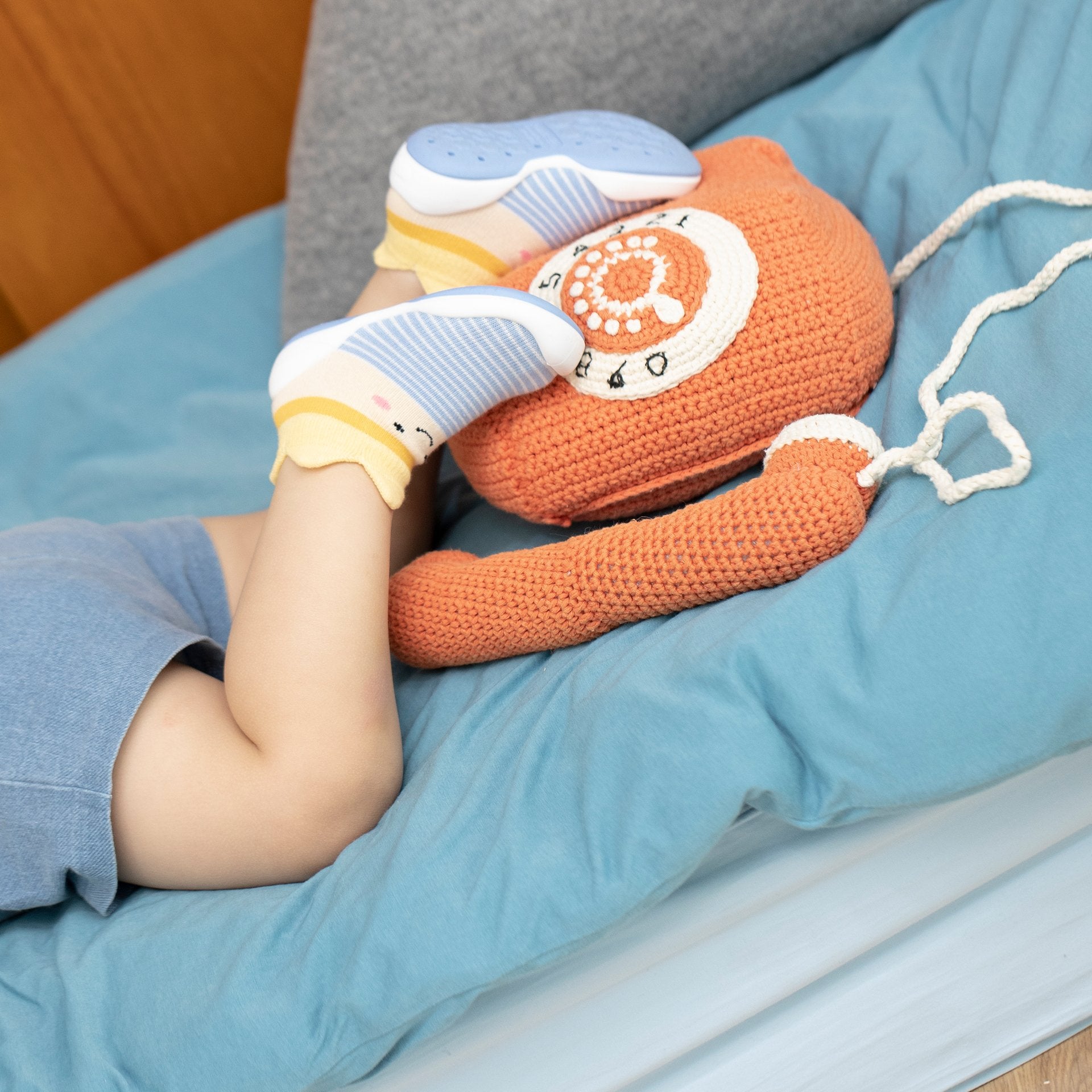 Child lying on a blue bed with feet propped up on an orange knitted telephone toy.