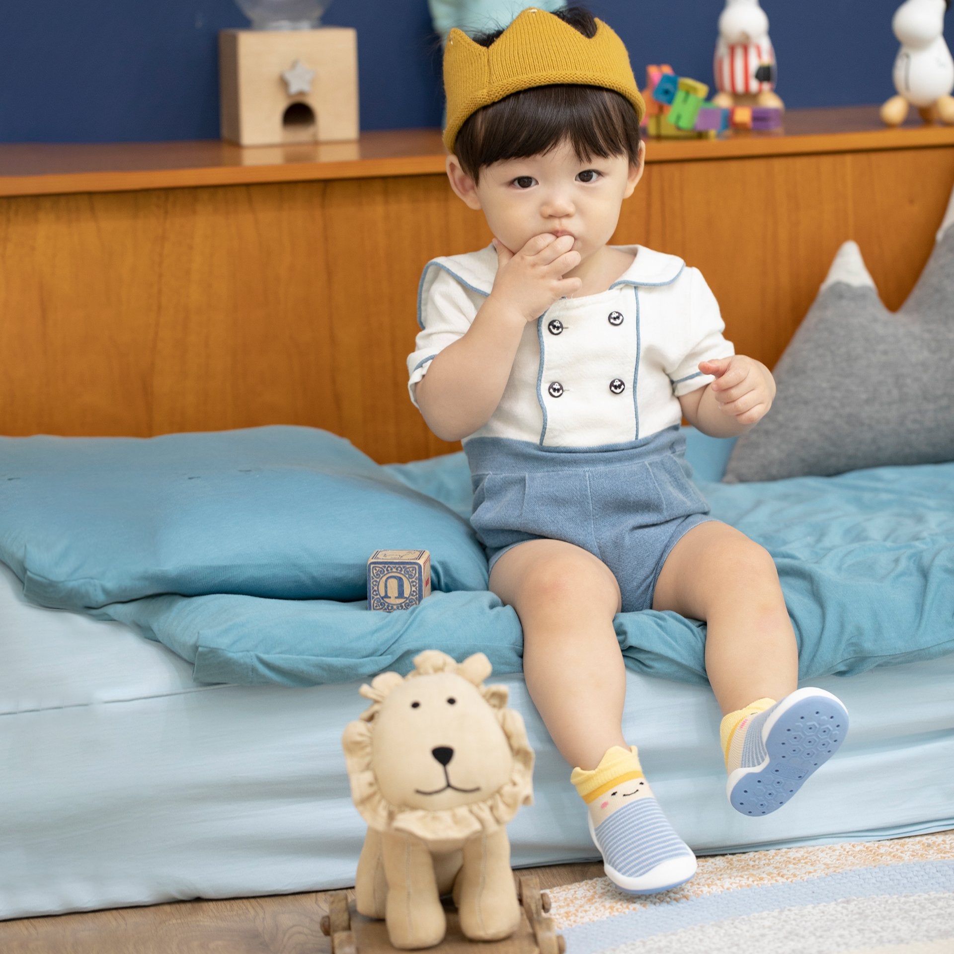 A child in a crown, blue romper, and striped socks sits on a blue bed next to a toy lion.