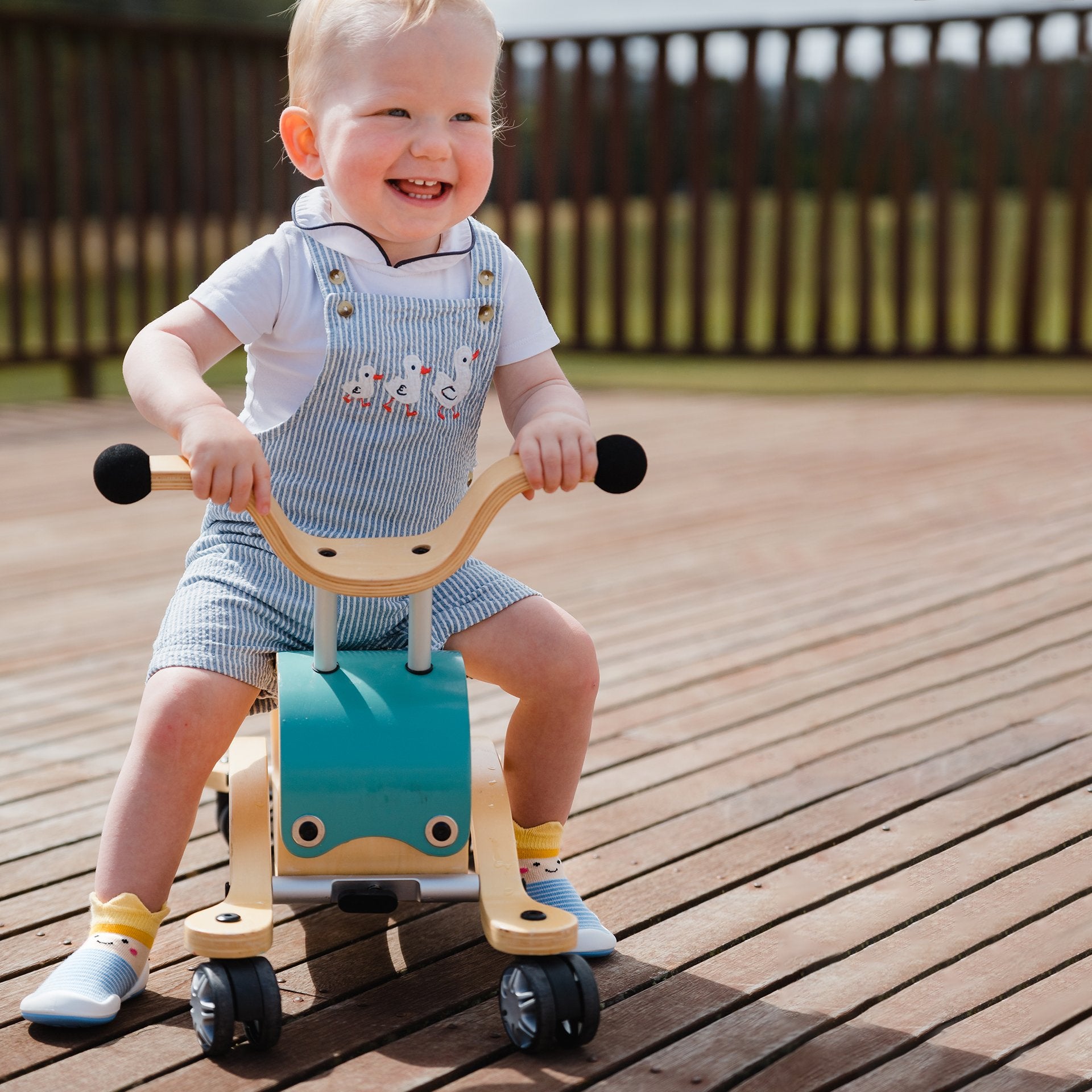 Smiling child on a teal and wooden ride-on toy. Light blue and white striped overalls with duck embroidery. Wooden deck.