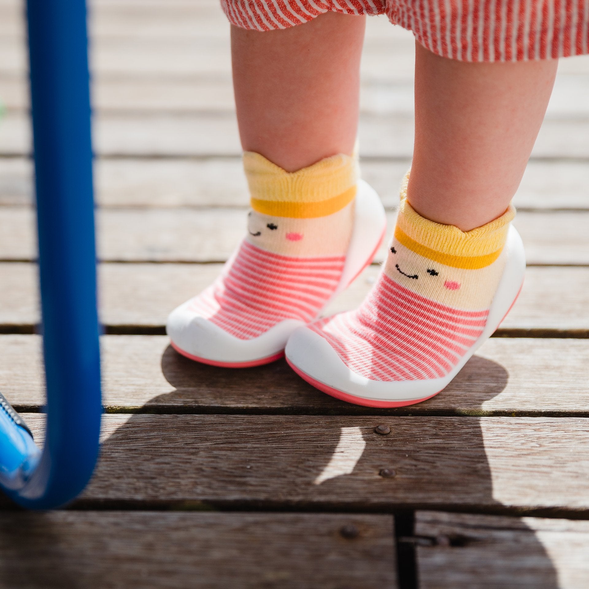 Toddler's feet wearing red and white striped socks with a yellow top and a smiling face. They are standing on a wooden deck.