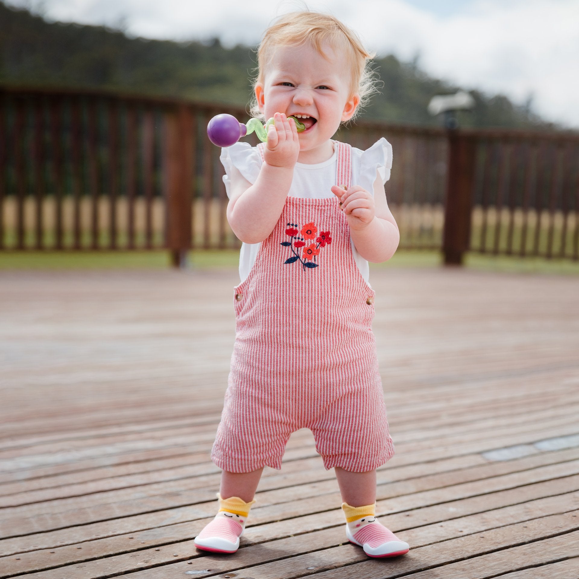 Baby in red and white striped overalls, biting on a purple and green toy, stands on a wooden deck.