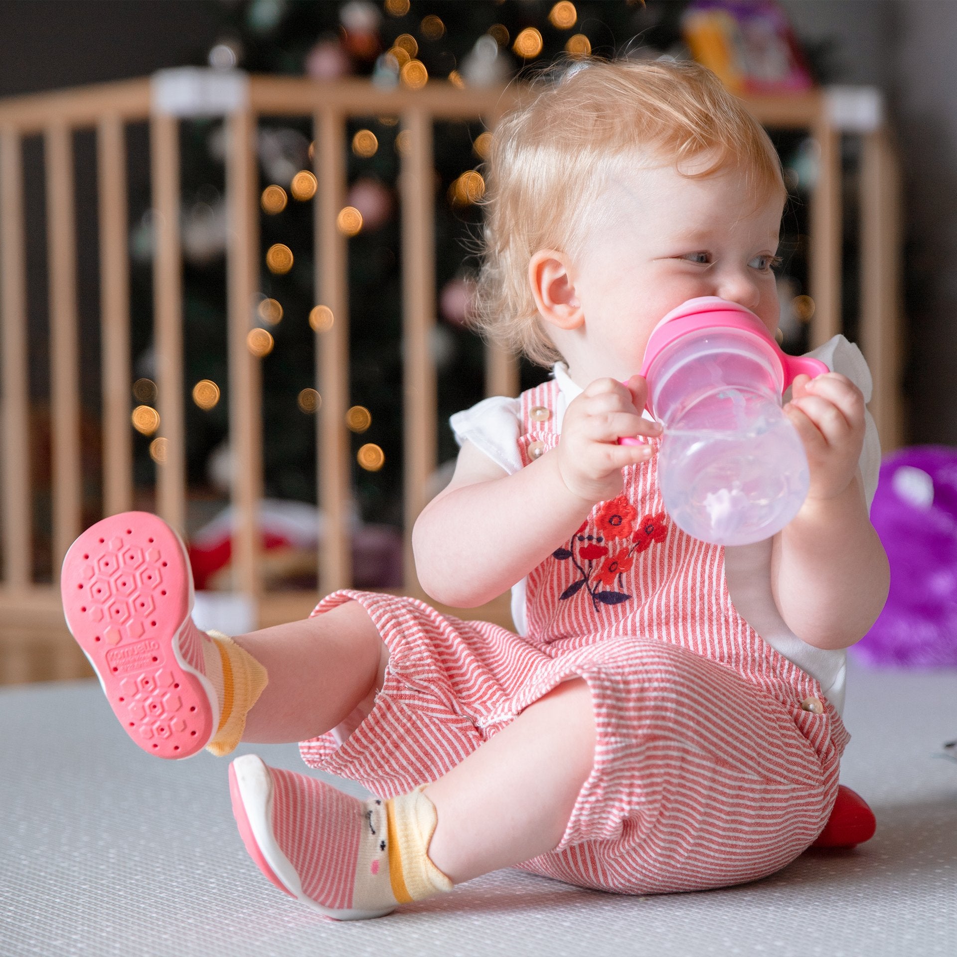 Baby drinking from a pink bottle while wearing striped overalls and socks.