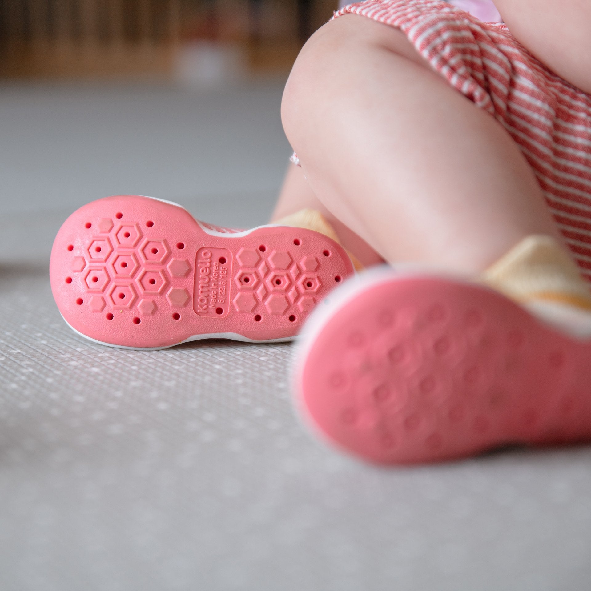 Baby shoes with pink soles, child's legs, striped dress, gray floor.