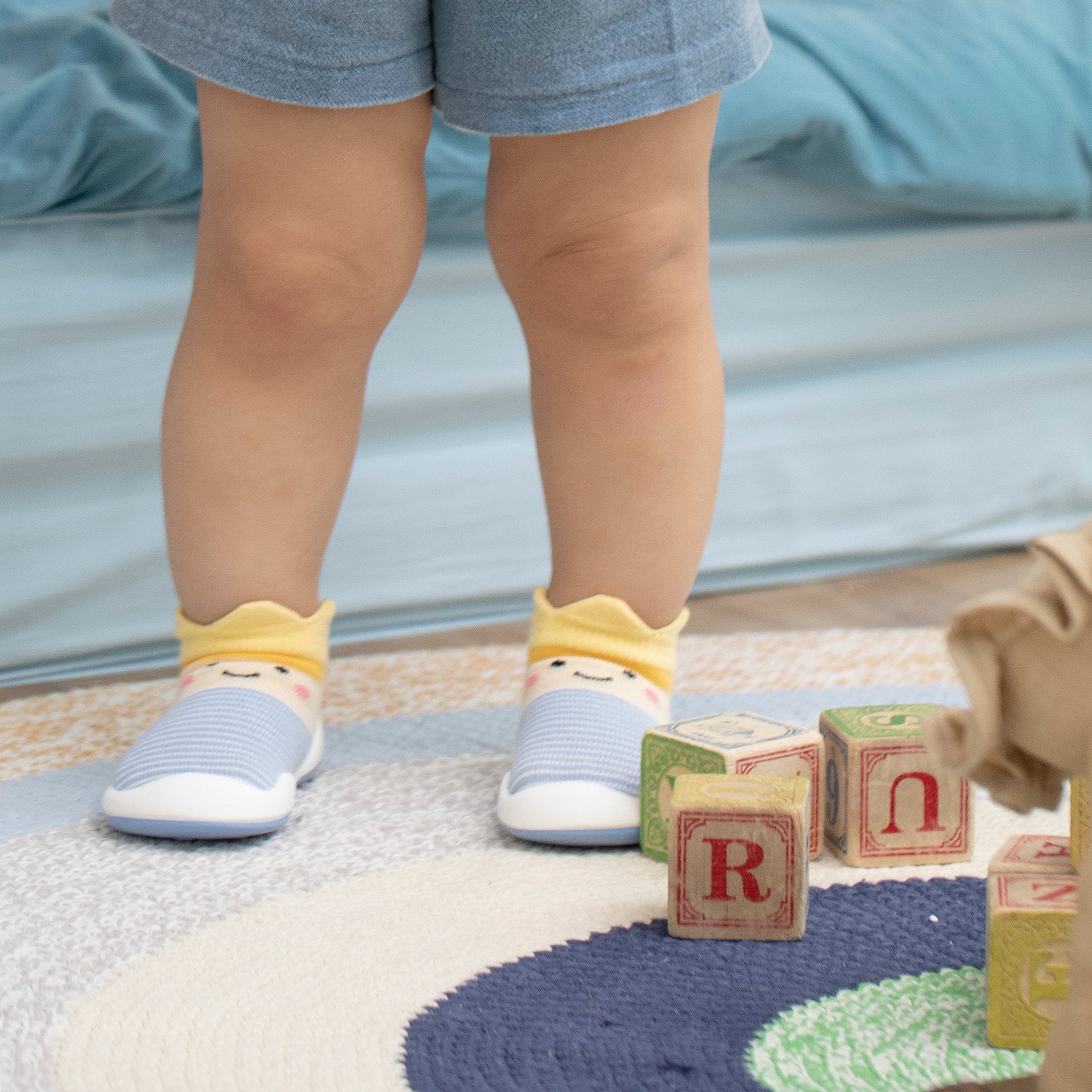 Child's legs and feet wearing blue and white striped socks, standing on a colorful rug with wooden alphabet blocks.