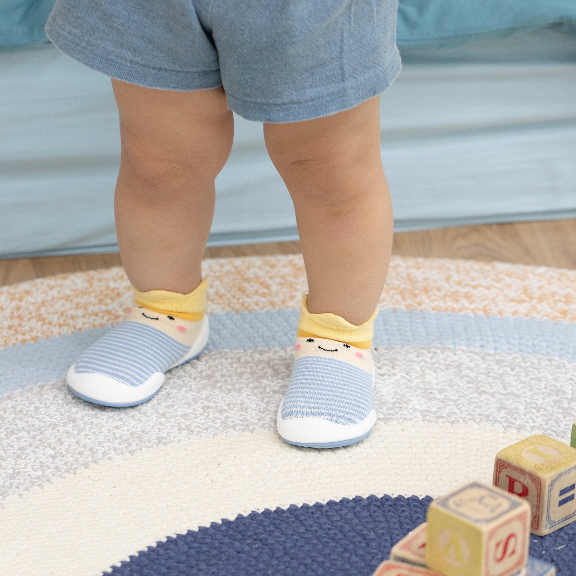 A child stands on a colorful rug in blue shorts and blue and white shoes with a smiling face design.