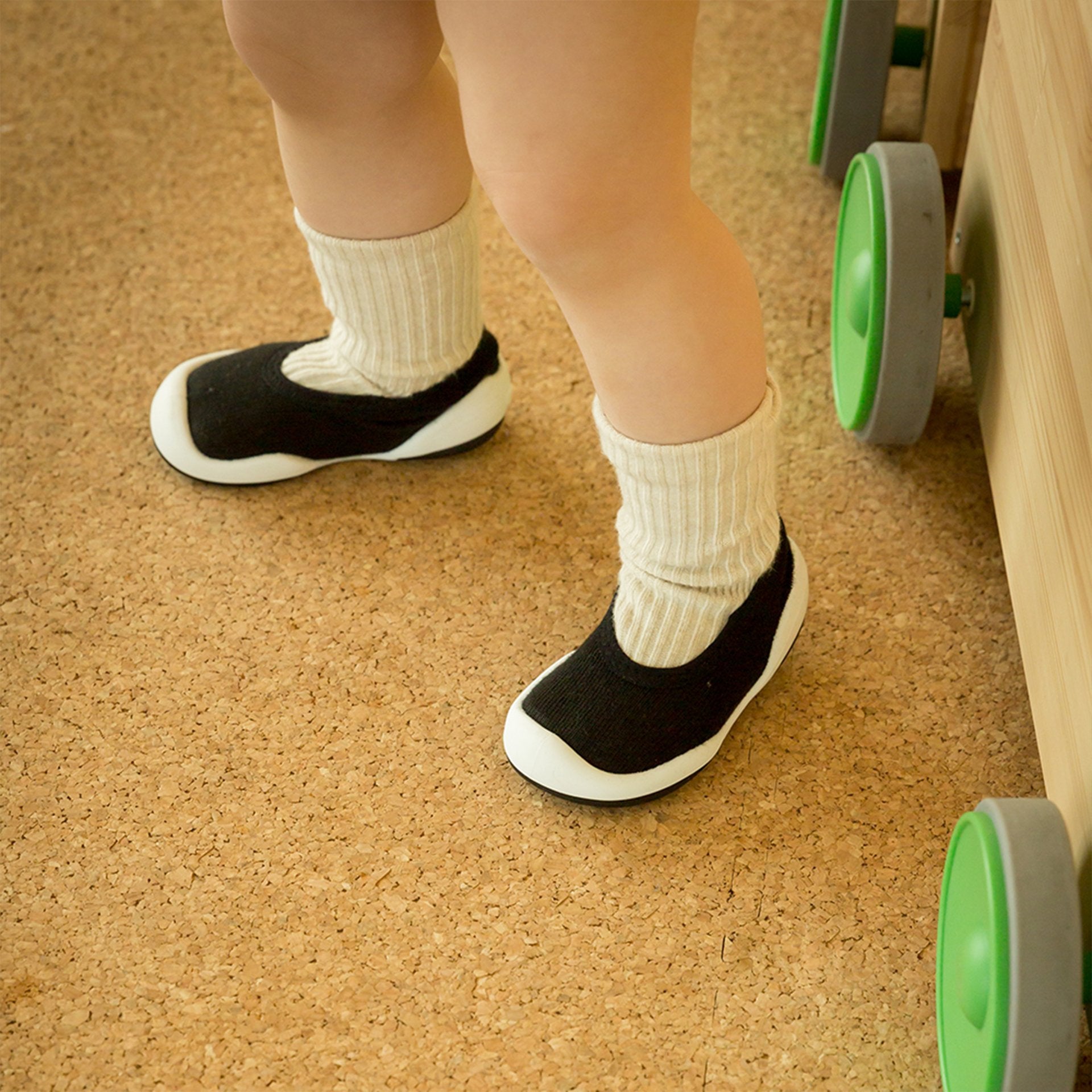 Toddler wearing cream socks and black slip-on shoes standing on a cork floor.