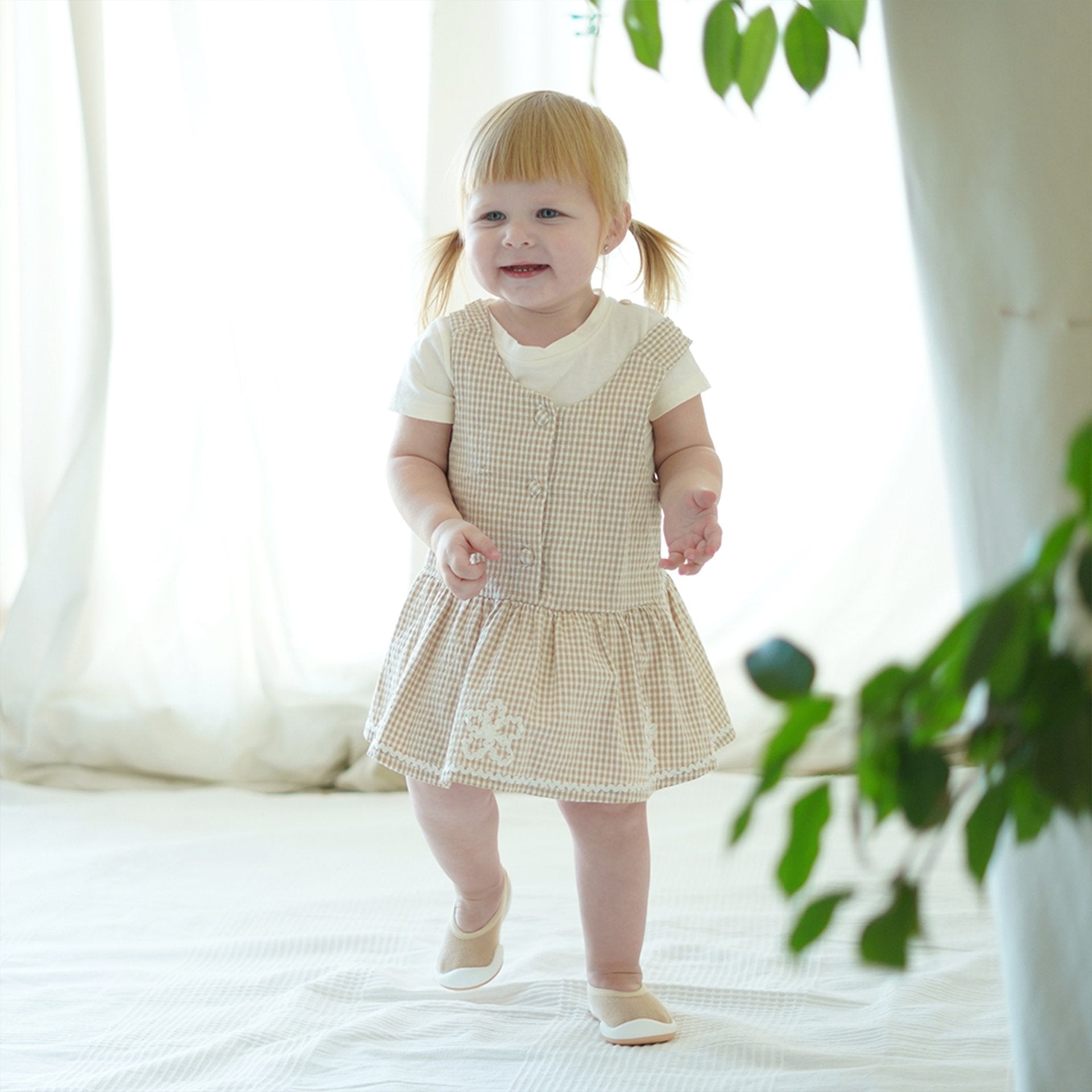 Toddler girl in beige checkered dress and matching shoes, smiling.