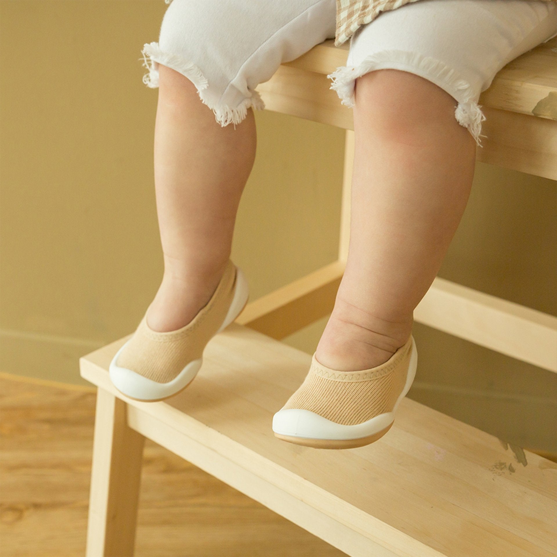 Toddler wearing beige and white shoes, fringed white pants, sitting on wooden steps.