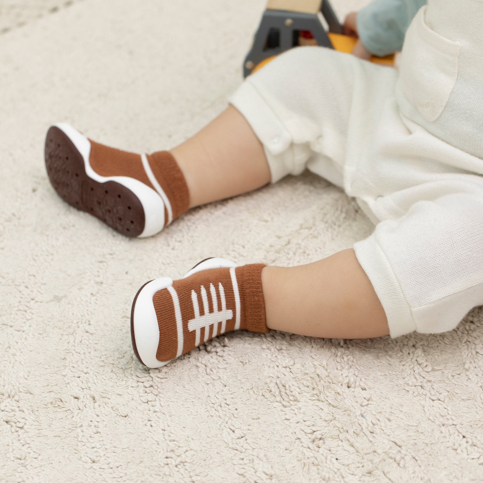 Child in white romper wearing brown football-themed sock shoes on carpet.