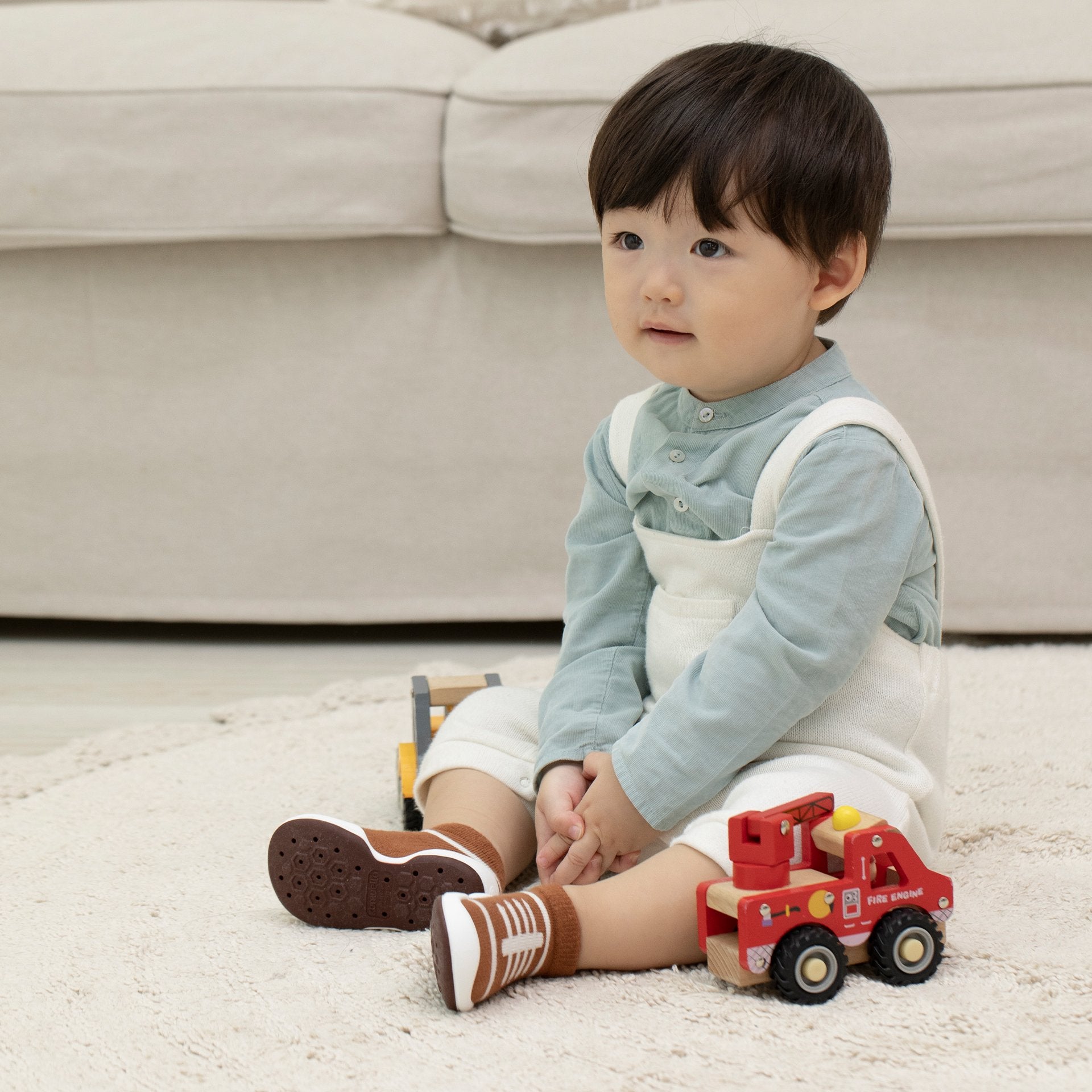 Child sitting on carpet with wooden toy vehicles, wearing blue shirt, white overalls, and brown-and-white socks.