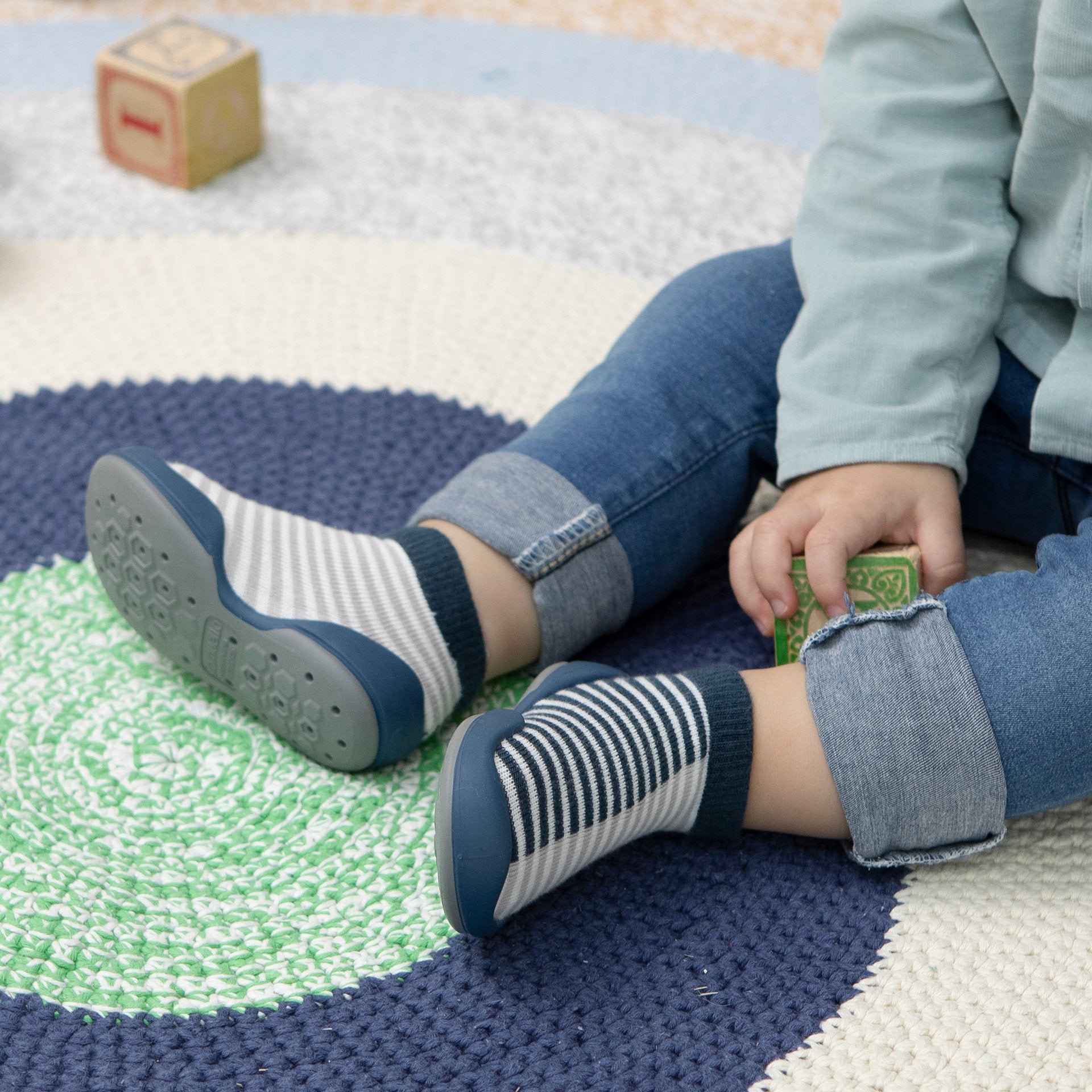 Toddler wearing blue and white striped slipper-socks, blue jeans, and light blue shirt while sitting on a colorful circular rug with building blocks.