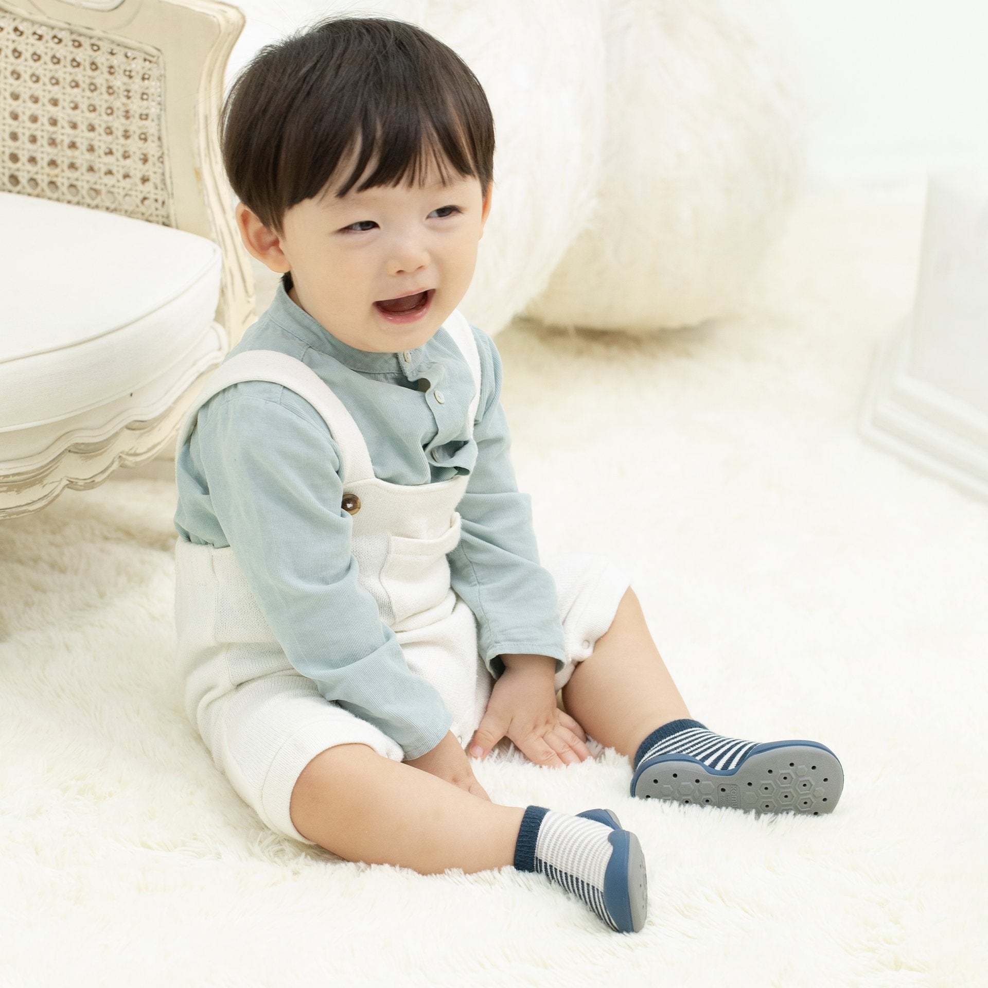 Baby sitting on a white rug, wearing blue shirt and white overalls.