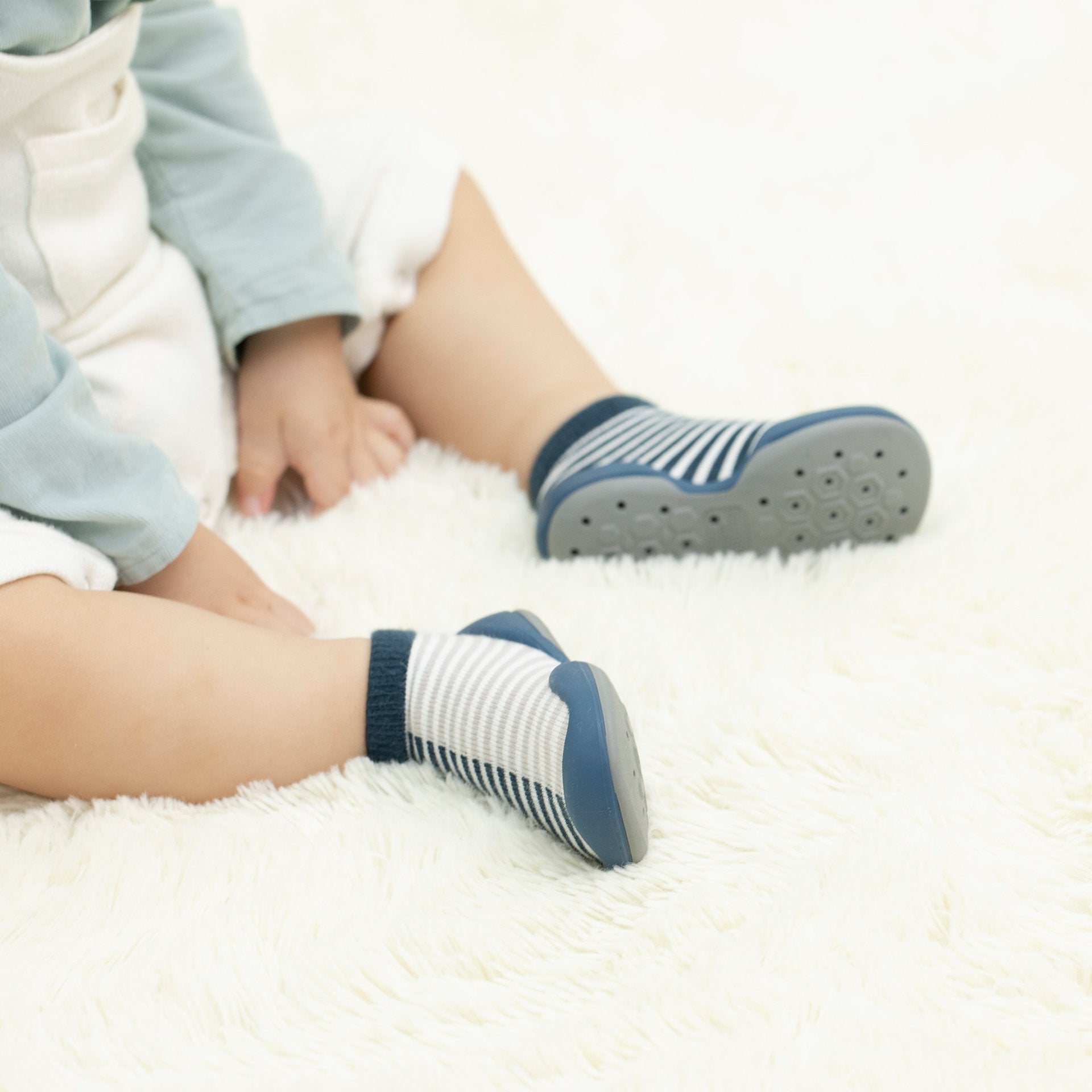 Baby sitting on white fur rug, wearing light blue top, white shorts, and striped socks with blue soles.