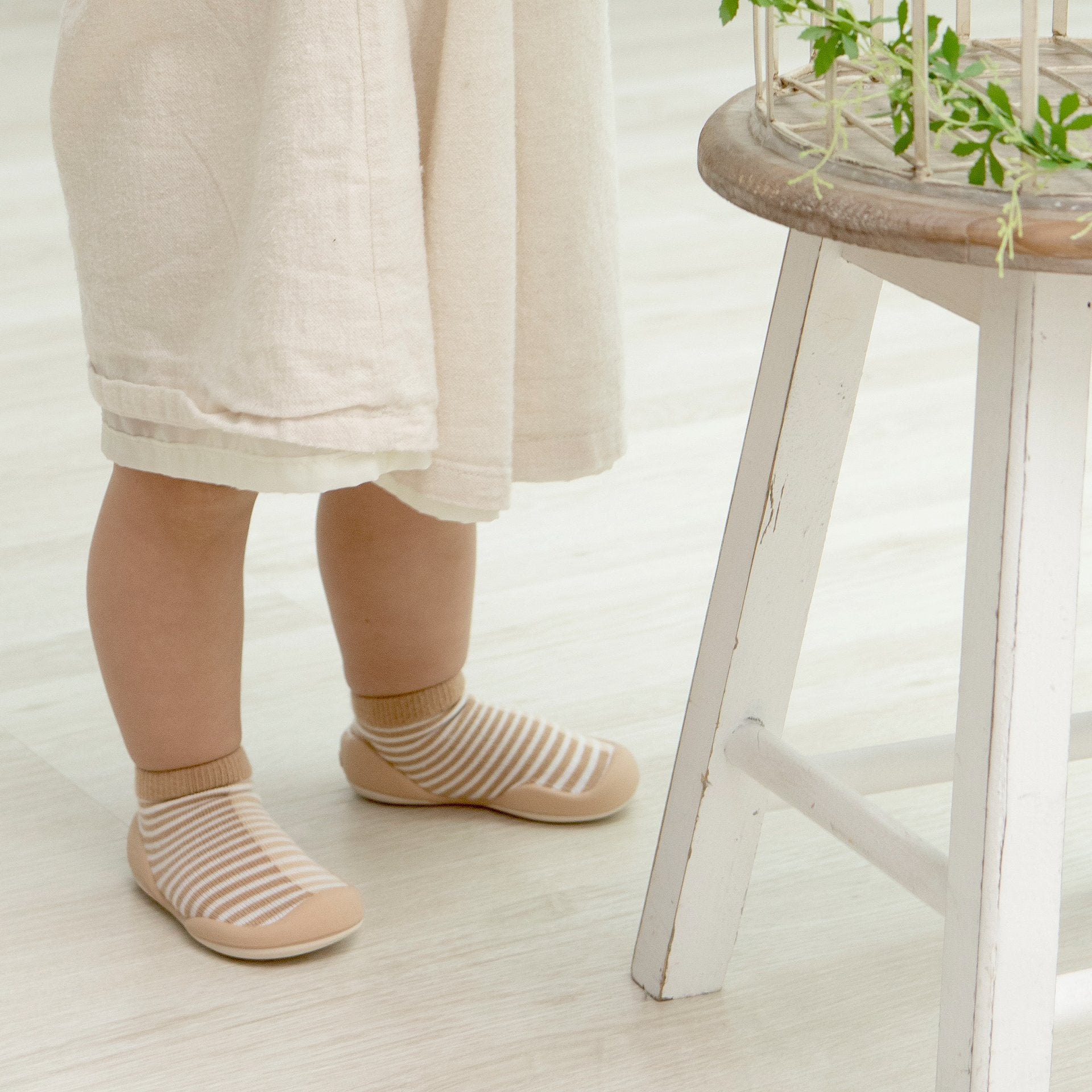 Baby in striped socks and white linen dress next to white stool.