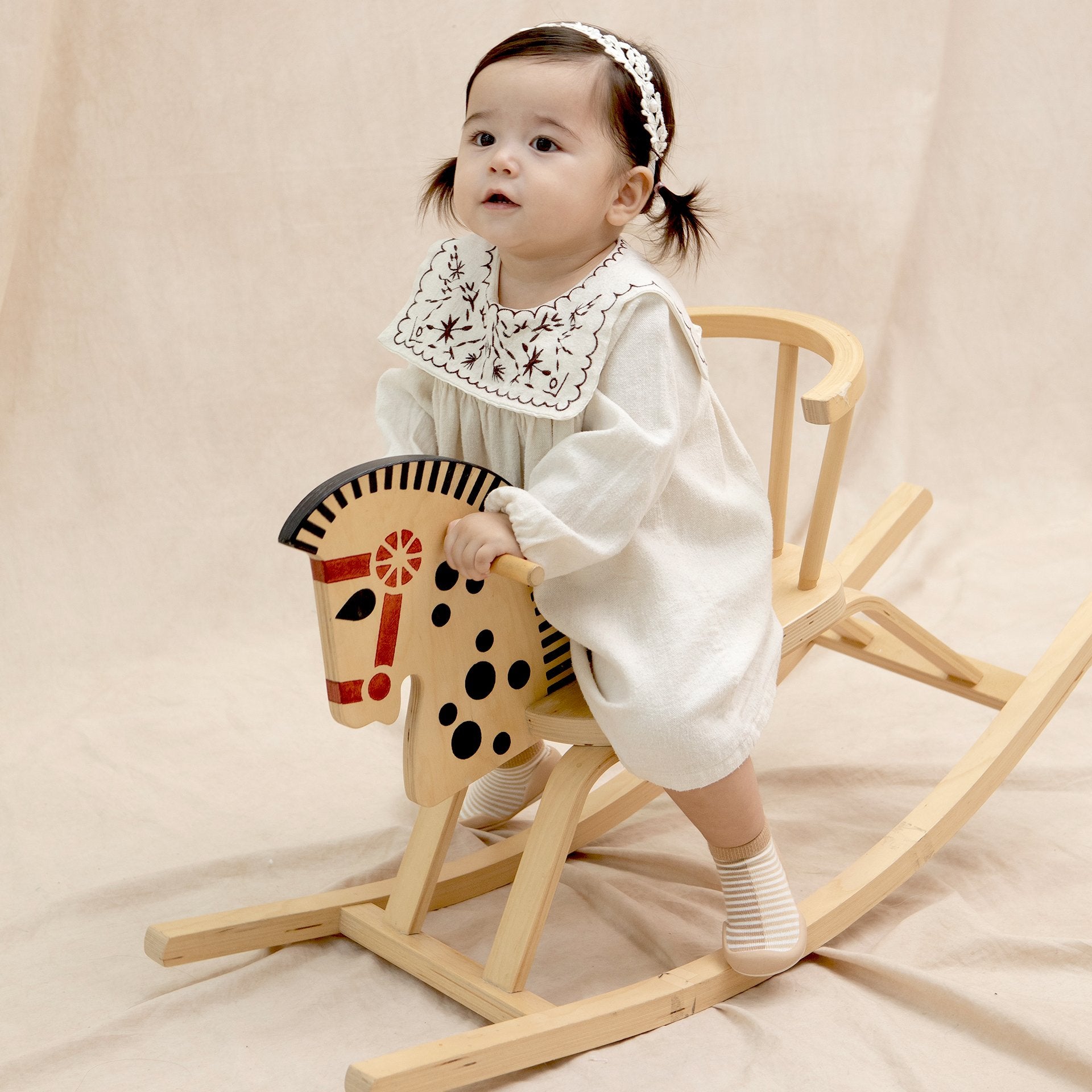 Baby girl on a wooden rocking horse, wearing a white dress with an embroidered collar and a white headband.