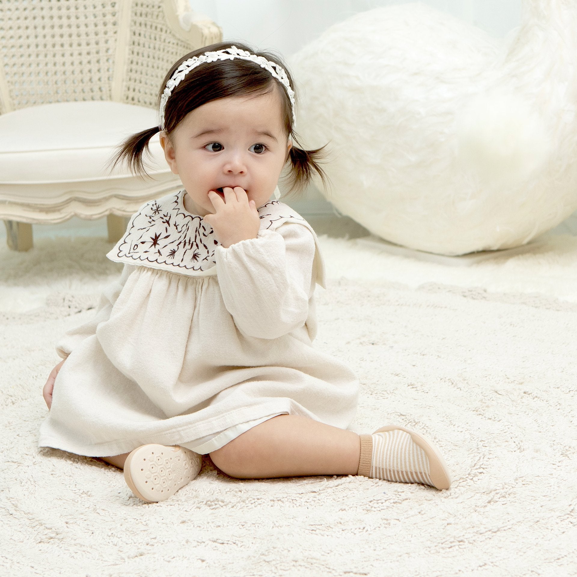 Baby in white dress with headband sitting on white carpet.