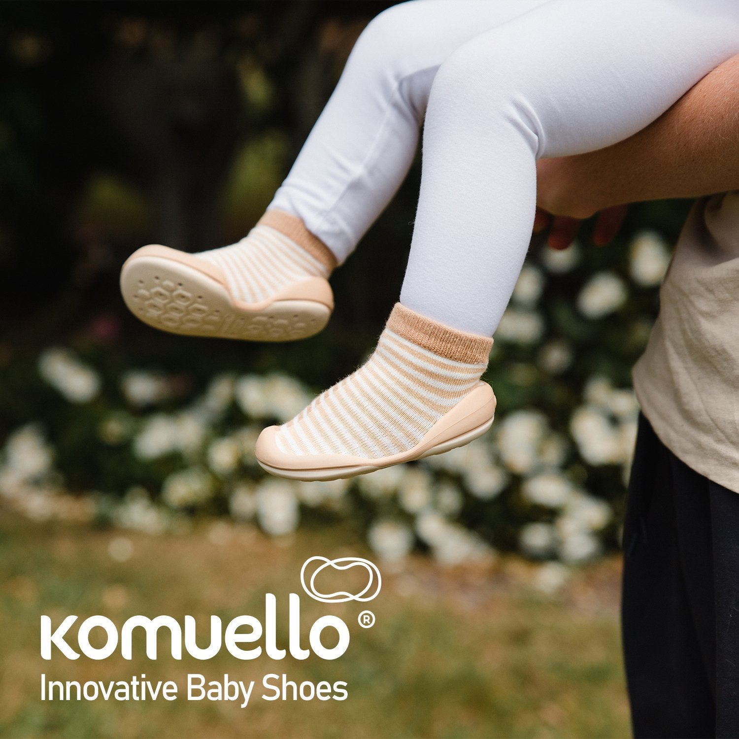Close-up of baby legs in white leggings and striped cream and tan Komuello baby shoes, held in someone's arms outdoors.