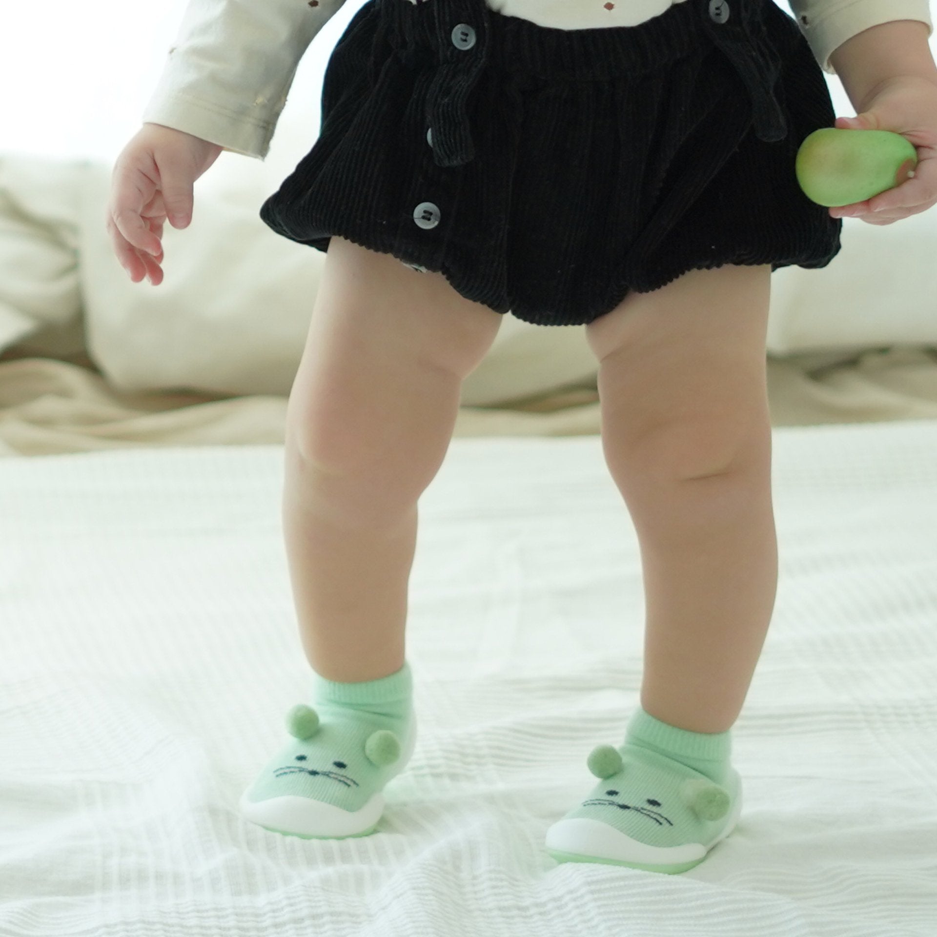 A baby stands on a white blanket wearing mint green cat-themed socks and a black corduroy skirt with buttons.